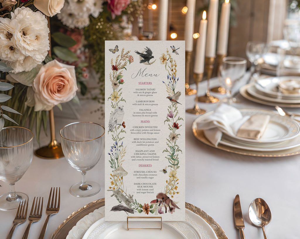 An enchanted forest menu with various woodland animals and bird graphics in a gold stand on a white plate sitting on a long white table