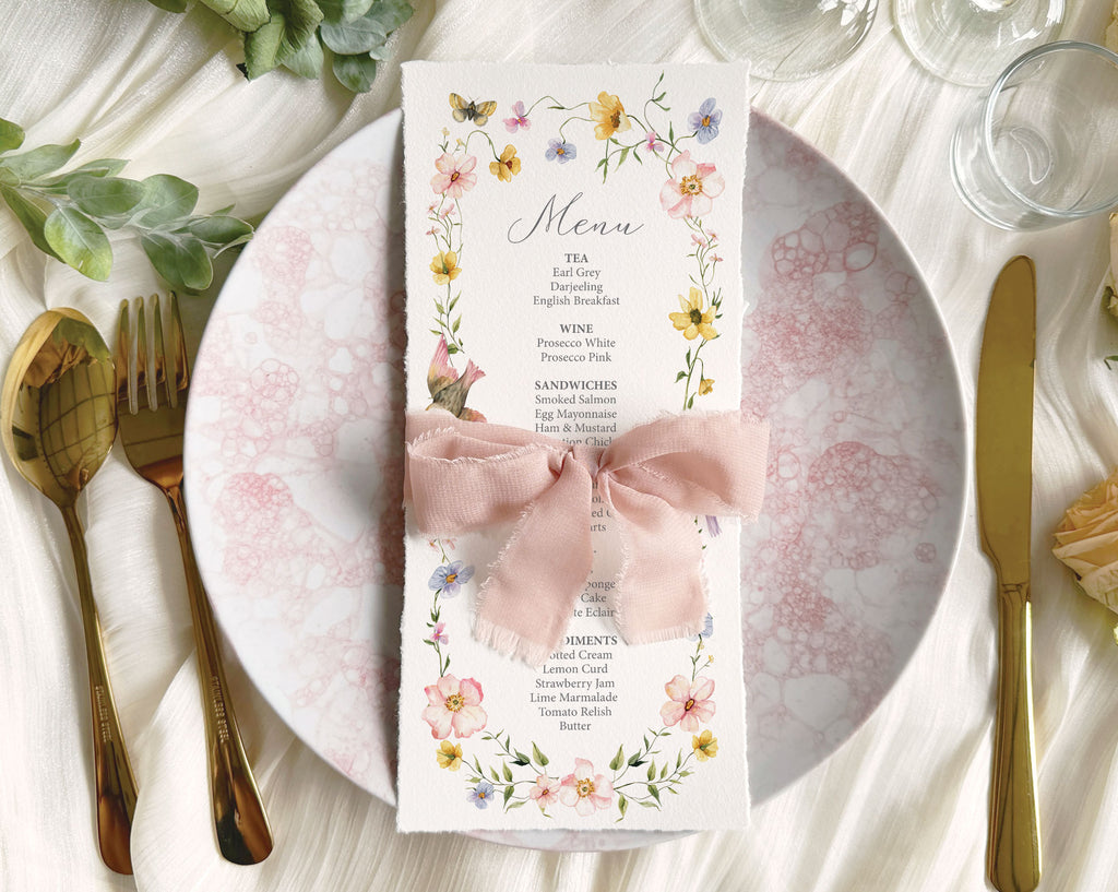 Menu card with floral design and birds, wrapped with a pink bow on a table setting with plates, cutlery, and glasses.