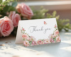 Floral 'Thank you' card with blush pink flowers and a butterfly design on a white surface with pink roses in the background.