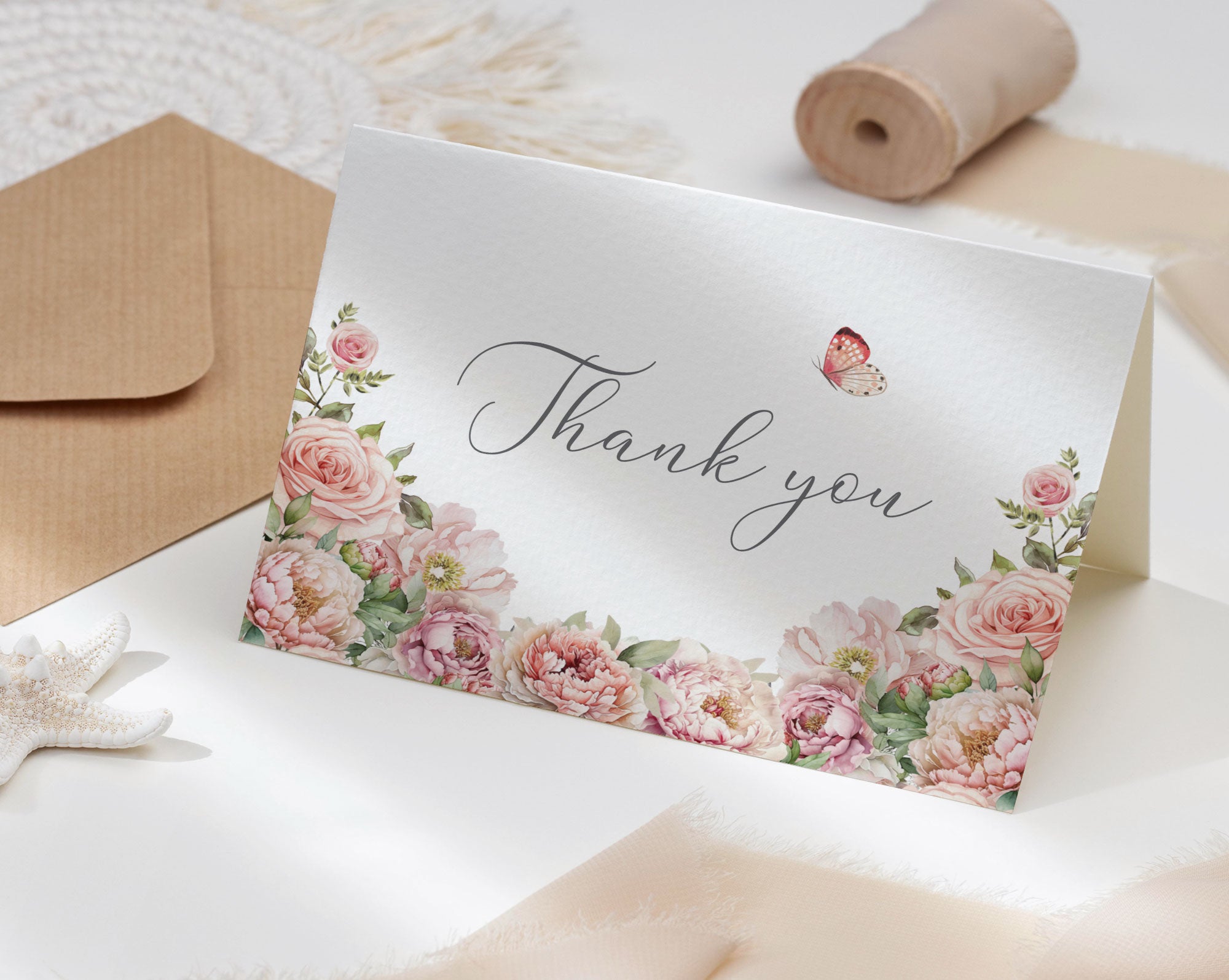 Floral 'Thank you' card on a white surface with a brown envelope and decorative ribbon roll.