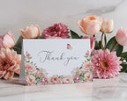 Thank You tent card with 'Thank you' text and butterfly design surrounded by pink flowers on a marble surface