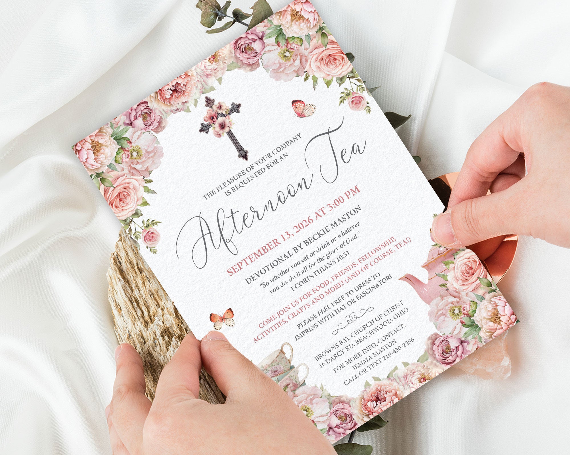 Invitation card with blush pink floral design and cross held by hands on a white fabric background