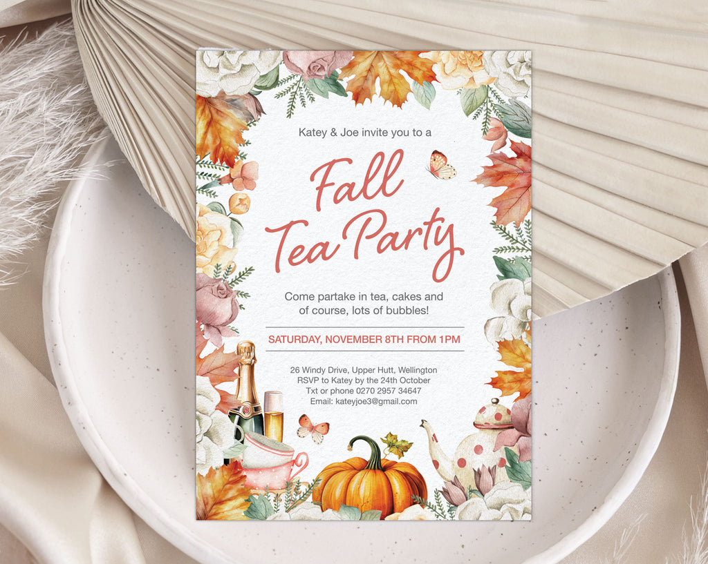 A vertical fall tea party invitation card with watercolor autumn leaves, pumpkins, teacups, and floral borders, styled on a white speckled plate with beige fabric and dried palm leaves.