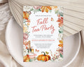 A vertical fall tea party invitation card with watercolor autumn leaves, pumpkins, teacups, and floral borders, styled on a white speckled plate with beige fabric and dried palm leaves.