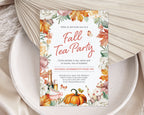 A vertical fall tea party invitation card with watercolor autumn leaves, pumpkins, teacups, and floral borders, styled on a white speckled plate with beige fabric and dried palm leaves.
