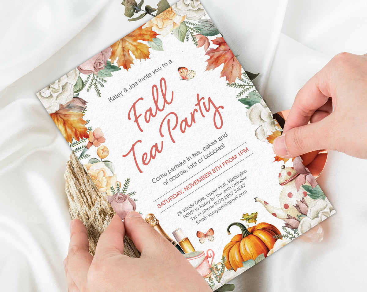 A person's hands holding a printed fall tea party invitation card over white fabric. The card features autumn watercolor illustrations including orange pumpkins, fall leaves, flowers, and a teapot, with the text 'Fall Tea Party' in large red script.