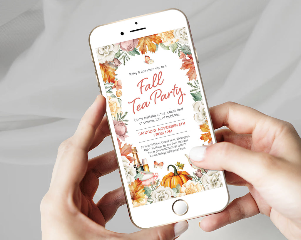 A person holds a white smartphone showing a fall-themed tea party invitation design with floral borders, pumpkins, and autumn leaves. The hands and phone are centered against a soft light gray fabric background.