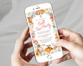 A person holds a white smartphone showing a fall-themed tea party invitation design with floral borders, pumpkins, and autumn leaves. The hands and phone are centered against a soft light gray fabric background.