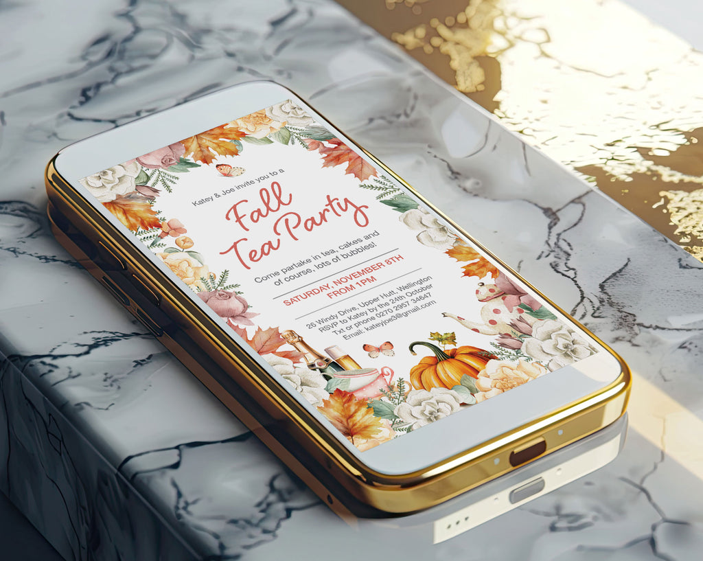 A gold-framed smartphone lying diagonally on a white and gray marble surface, displaying an autumn-themed tea party invitation design with fall leaves, pumpkins, flowers, and teacups.