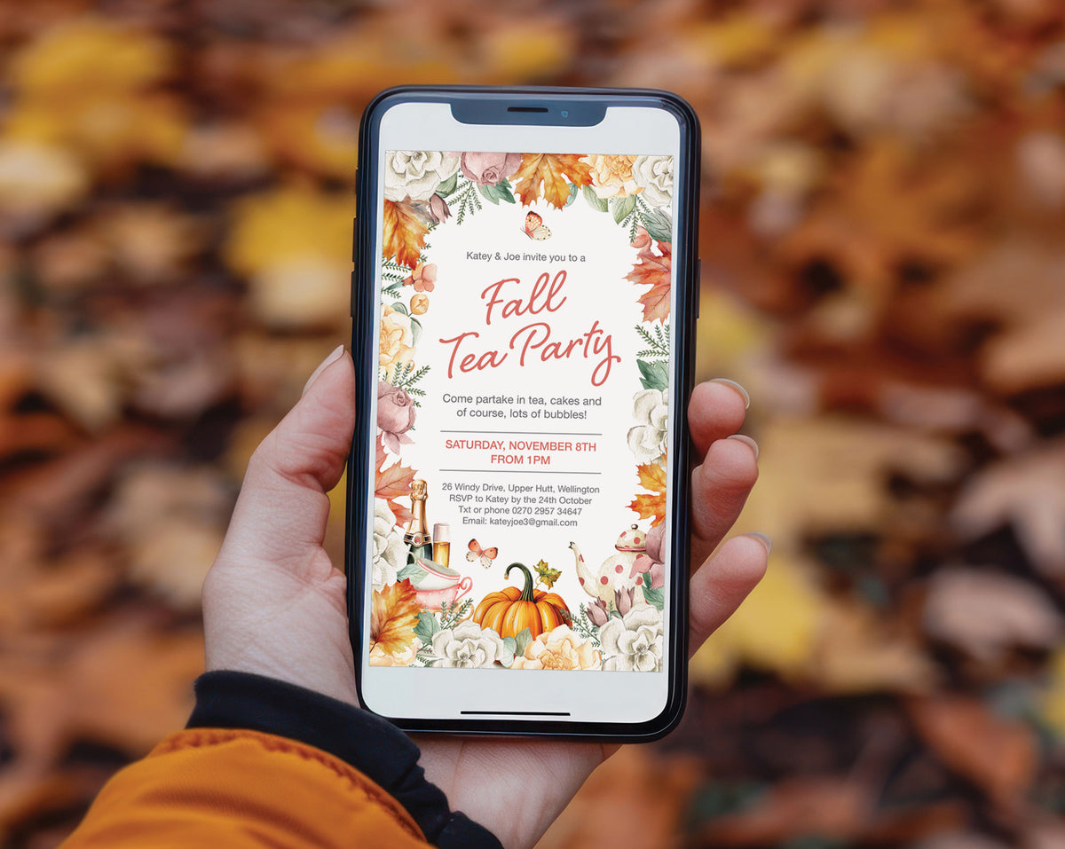 A hand holding a smartphone that displays a fall-themed digital invitation template with autumn leaves, pumpkins, and floral designs, set against a blurred background of real fallen leaves.