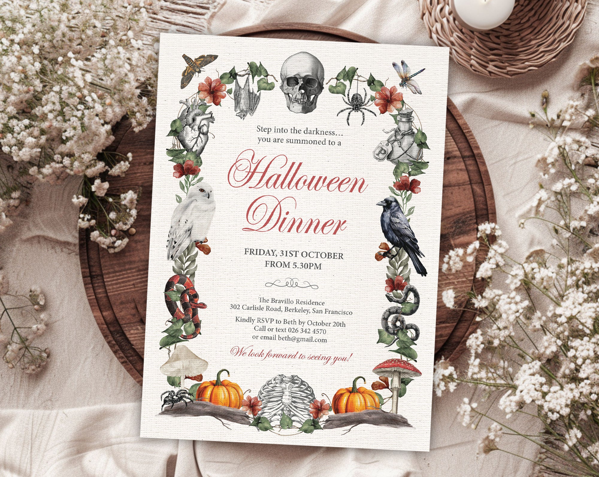 A Halloween dinner invitation card styled in a flat lay on a beige fabric surface with white baby's breath flowers, a woven tray, and a candle. The card features a decorative botanical border with a skull, crows, spiders, snakes, pumpkins, and autumn leaves, with the text 'Halloween Dinner' in red script.