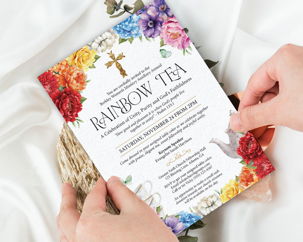 two hands holding a rainbow tea church event invitation with bright flowers, gold cross and teapot, with a white silk cloth in the background