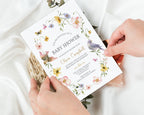 Baby shower invitation with floral and bird design held by hands on a light background