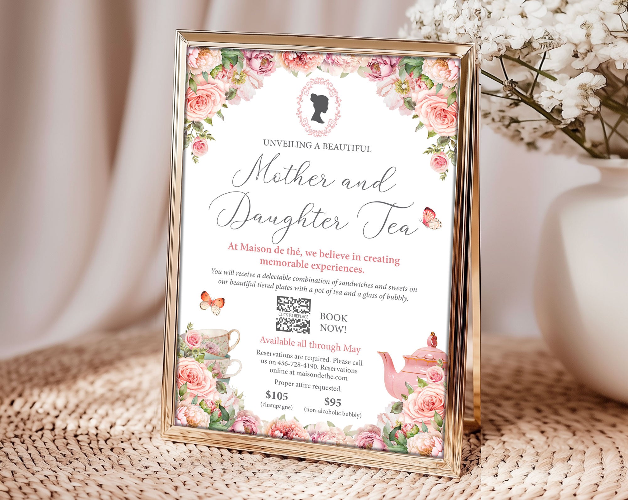a gold frame with a mother and daughter tea invitation with cameo and QR code showing on a rattan mat next to a vase of white flowers