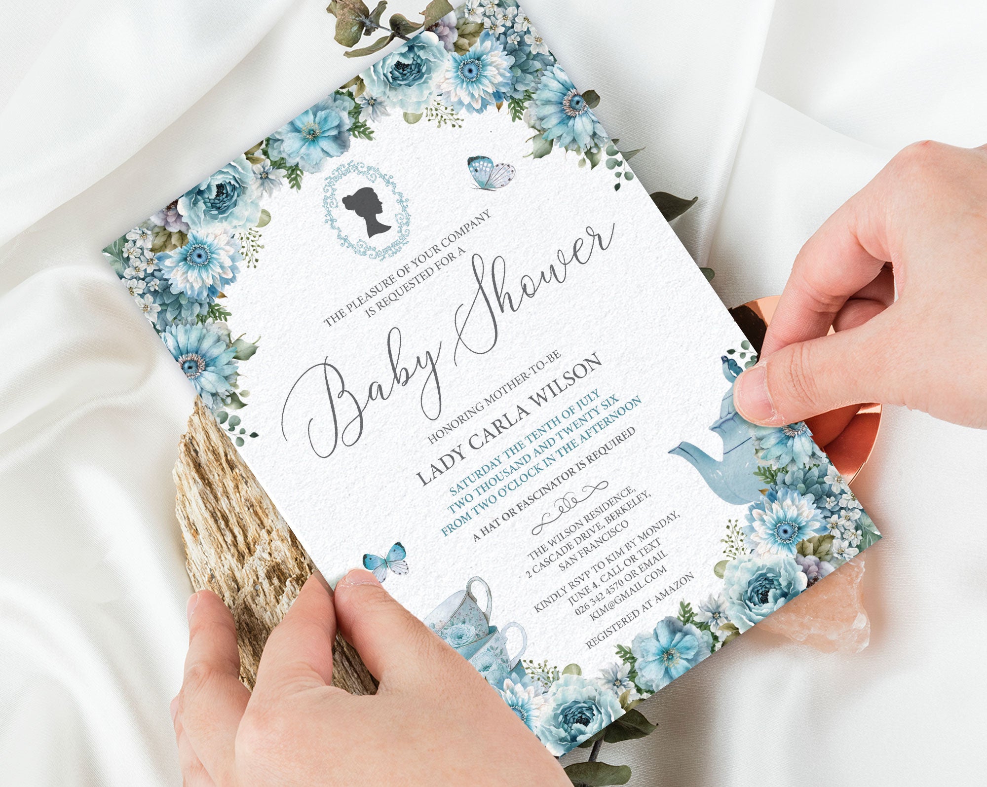 two hands holding a 5 x 7 dusty blue regency style baby shower tea invitation over a white silk background