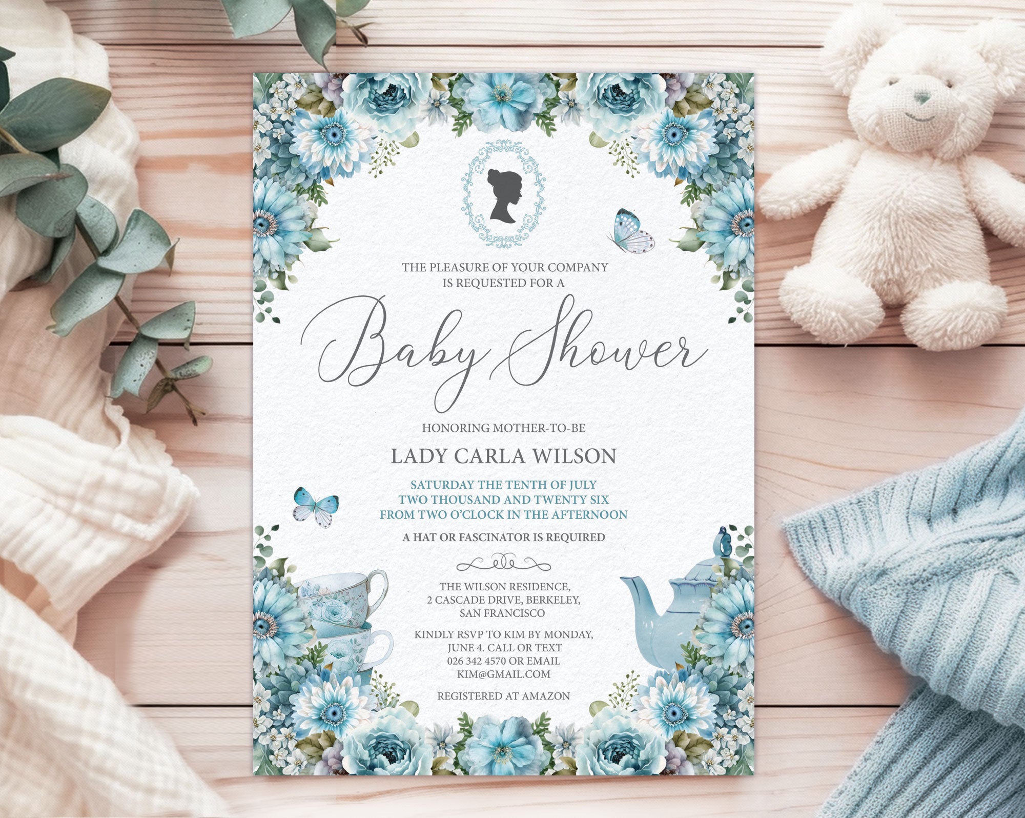 an elegant blue baby shower tea regency style invitation laying on the pink pattern back of the invite, on a white cloth with a white teddy bear and a wrapped gift