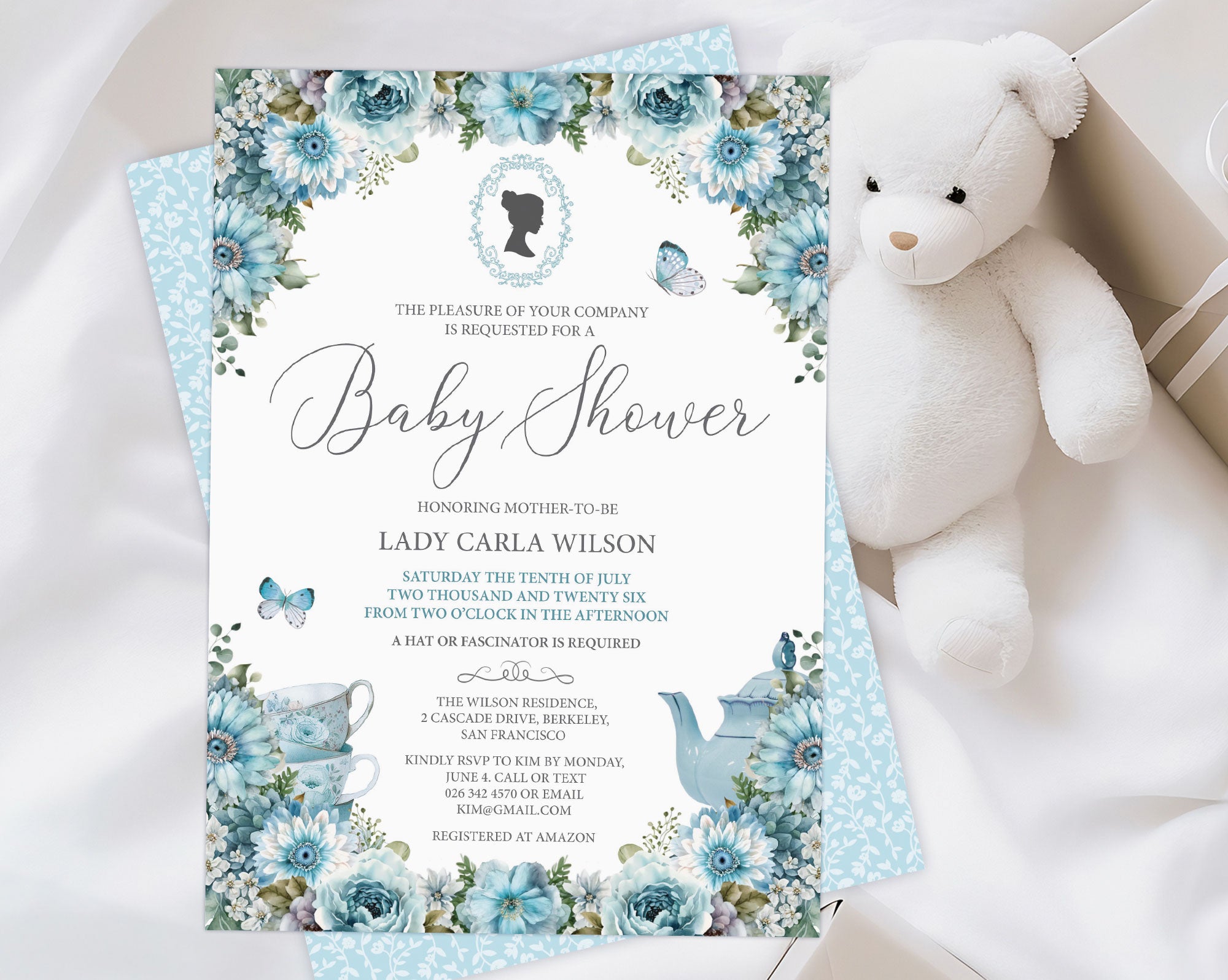 a dusty blue flower baby shower tea invitation with cameo and teapot, laying on a wooden background next to a white teddy bear, some baby blankets and eucalyptus