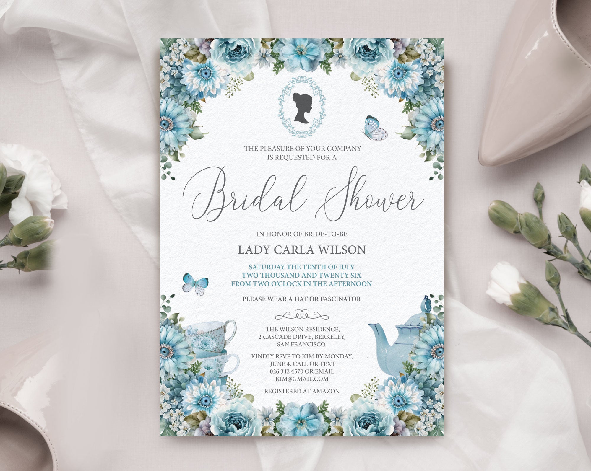 Pretty dusty blue flower bridal shower invitation laying on a beige silk background with a wedding shoe and some flowers