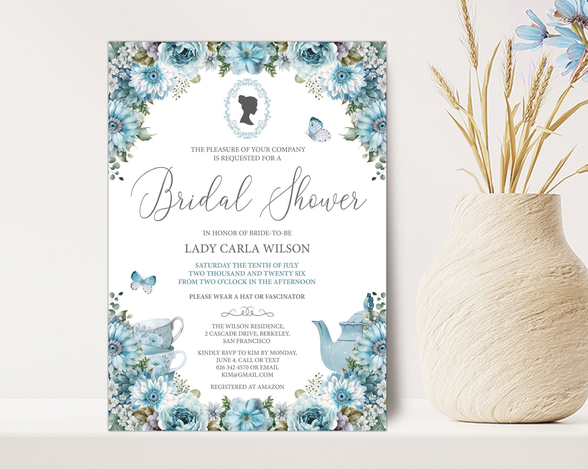 a 5 x 7 blue flower bridal shower invitation with victorian cameo, leaning up against a beige wall next to a vase full of dried blue flowers