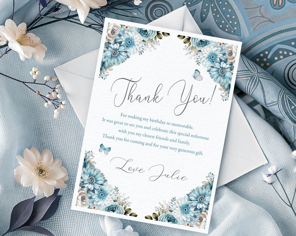 Elegant mockup of a thank you note with dusty blue flowers sitting on a white envelope next to white silk and batik cloth