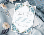 Elegant mockup of a thank you note with dusty blue flowers sitting on a white envelope next to white silk and batik cloth