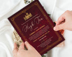 two hands holding a downton high tea for a birthday invitation with burgundy and gold art deco style design in front of a white silk background