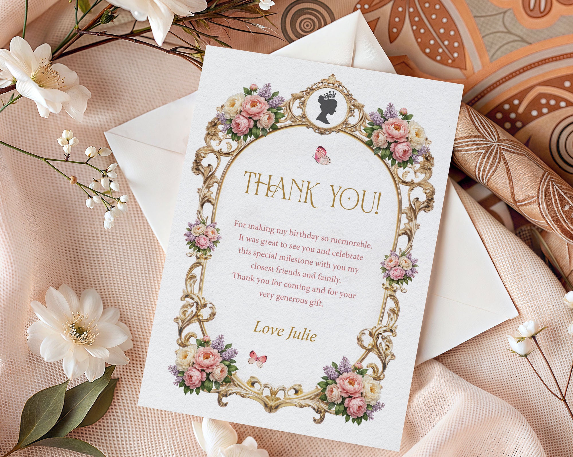 Elegant mockup of a royal thank you note with pastel peony flowers, gold frame and cameo, laying on a white envelope next to white silk and batik cloth