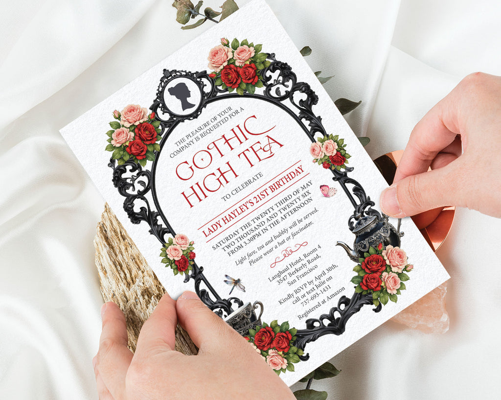 two hands holding a regency gothic high tea invitation with red and pink flowers, ornate black frame, cameo, teapot and cups with a white silk background behind