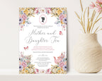 a 5 x 7 mother and daughter tea invitation standing up against a beige wall next to a vase of dried flowers