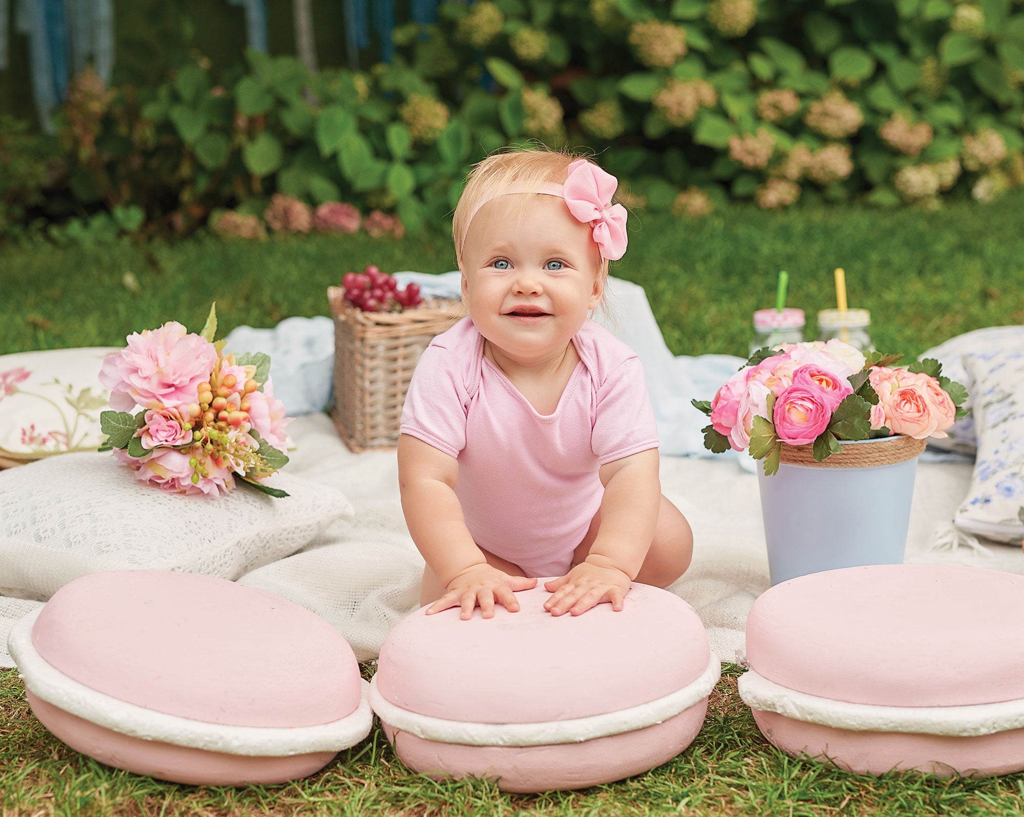 Baby in a pink outfit sitting on large pink macaron-shaped cushions outdoors with flowers and a blanket.