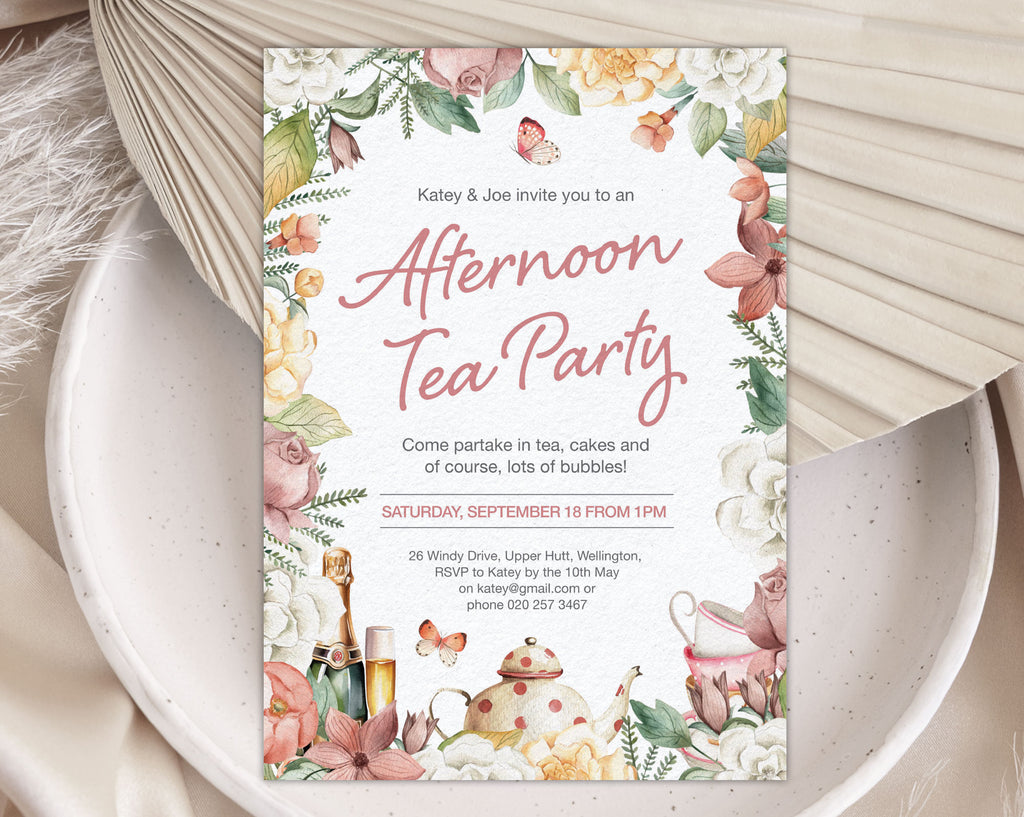 Floral afternoon tea party invitation with teapot and champagne graphics laying on a white plate with a dried leaf in the background