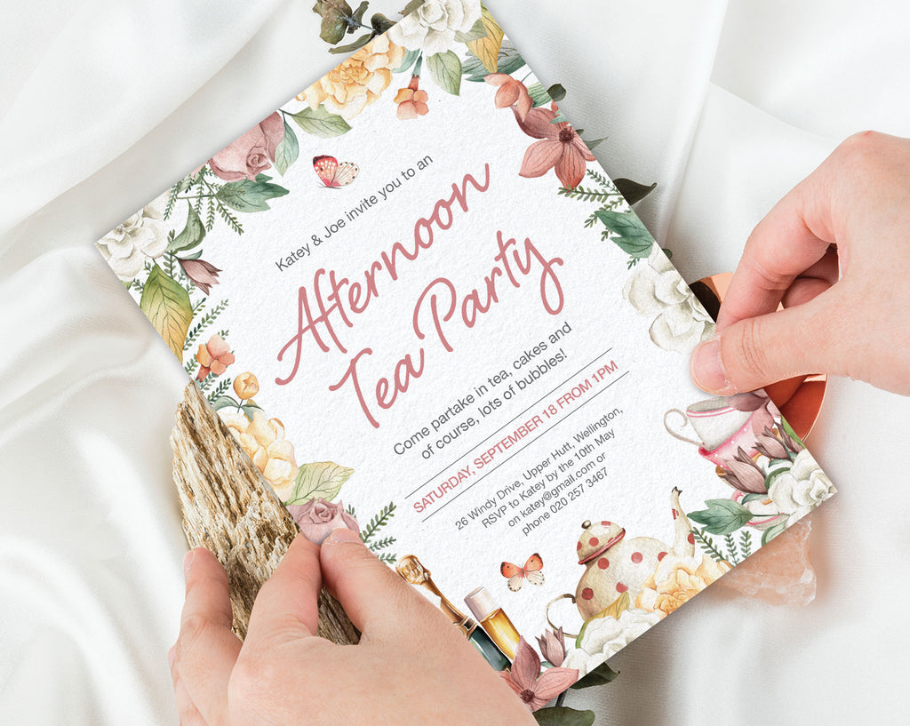 two hands holding an afternoon tea party invitation with a floral border, teapot, champagne and butterfly graphics over a white silk background