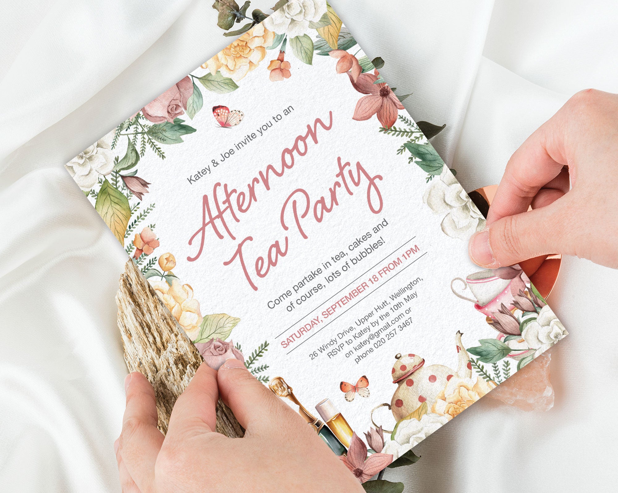 two hands holding an afternoon tea party invitation with a floral border, teapot, champagne and butterfly graphics over a white silk background