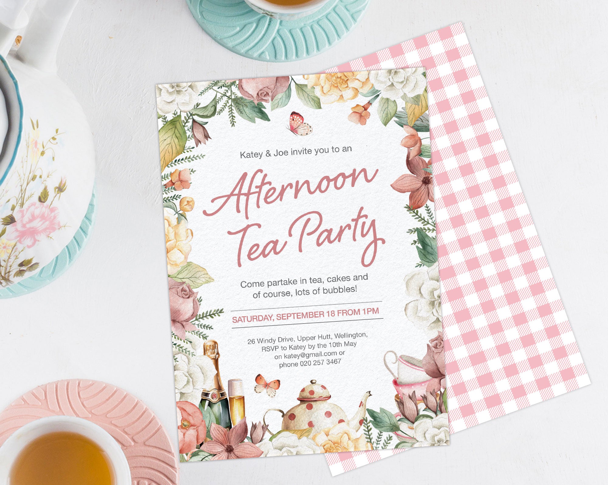 afternoon tea party invitation with floral border, showing the pink gingham back, laying on a white background next to a pastel tea set