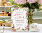 pretty tea table with an afternoon tea party invitation leaning up against a tiered food stand next to a vase of pink roses and a tea cup
