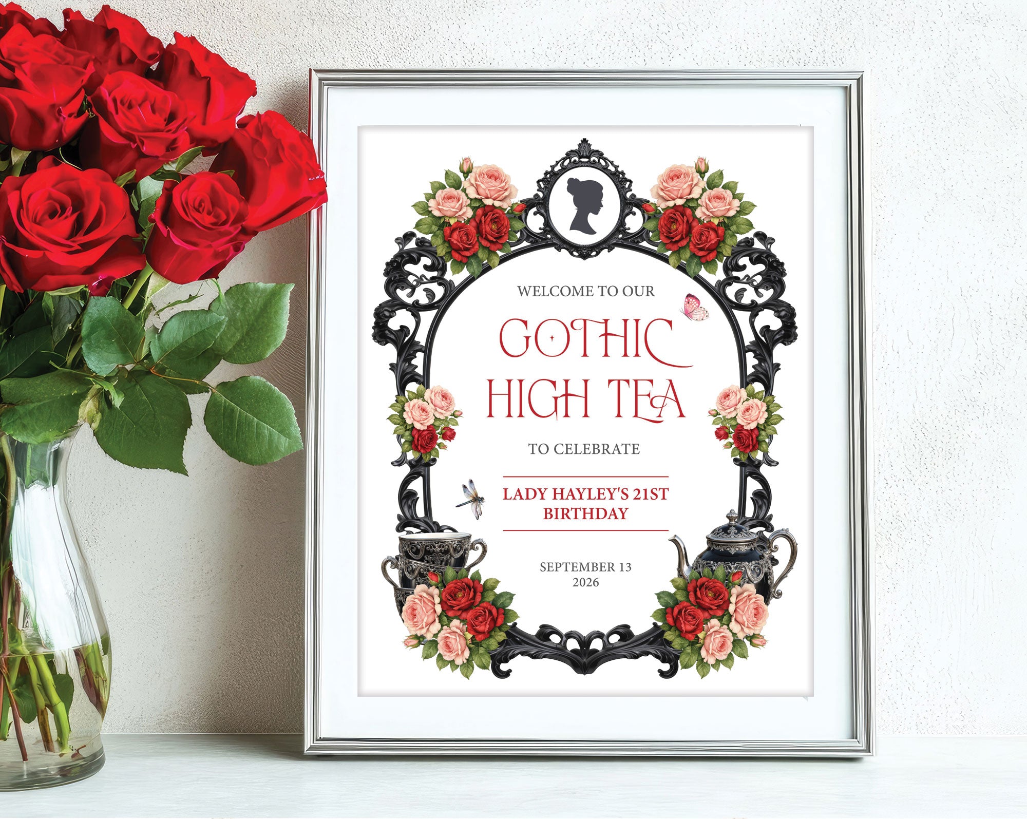 red and black gothic high small welcome sign in a silver frame leaning up against a white wall next to some red roses