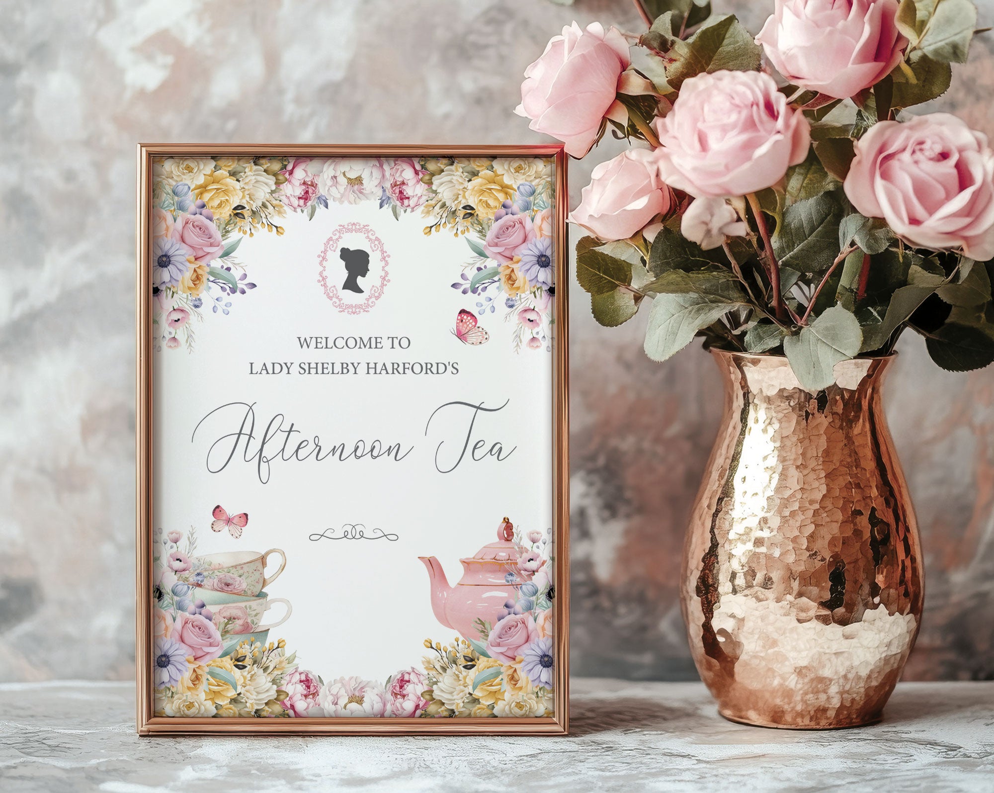 A gold framed welcome sign with pastel flowers, regency cameo and butterfly graphics, leaning up against a grey pattern wall next to a copper vase with pink roses.