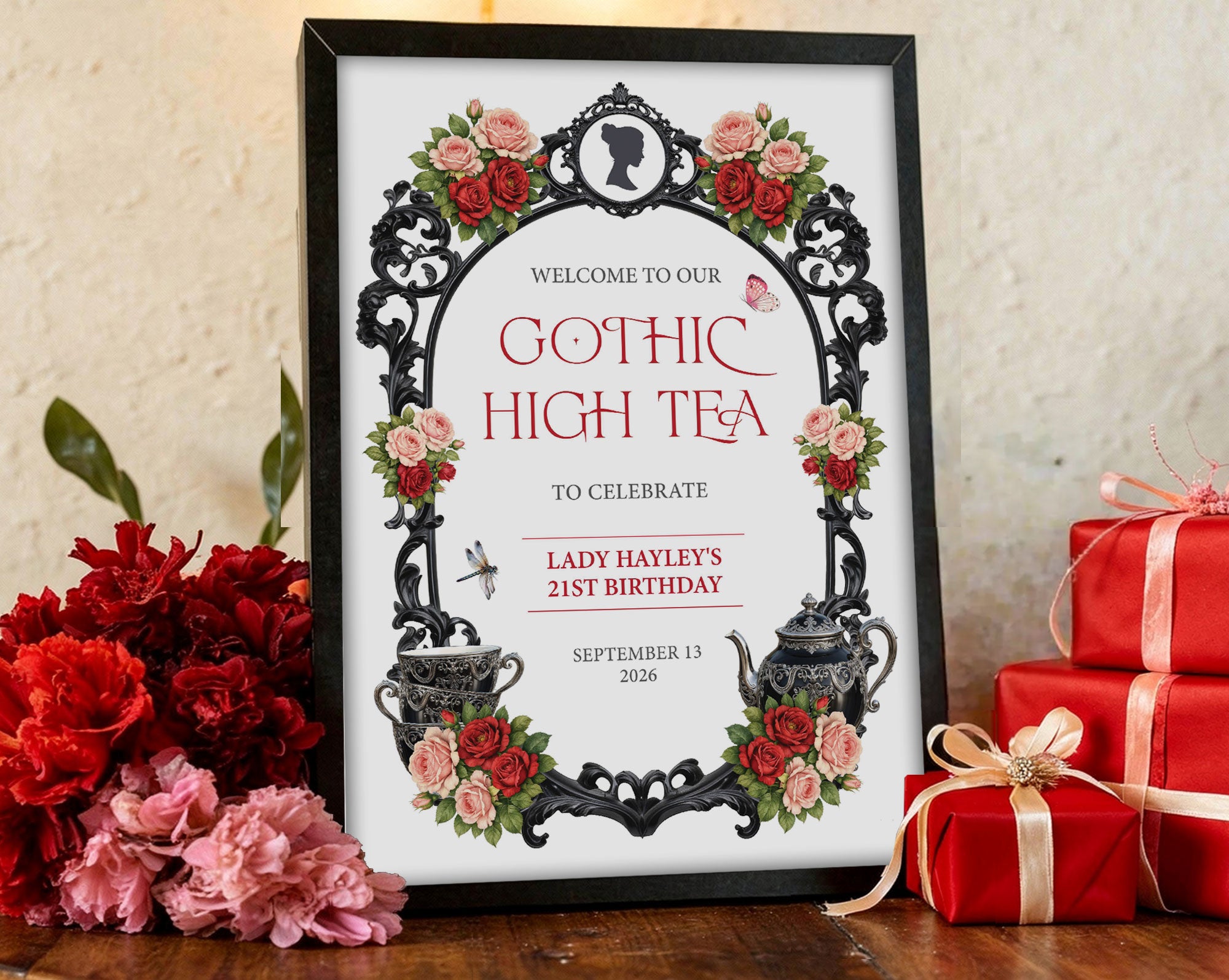 12 x 18 gothic high tea welcome sign with ornate frame, teapot, cameo and butterflies in a black frame leaning against a beige wall next to red packaged gifts and red and pink flowers
