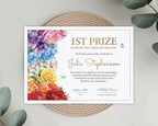 A printed A4 certificate for 1st Prize Rainbow Tea Table Decoration, featuring colorful watercolor floral illustrations on the left side and elegant gold typography, displayed on a woven placemat with eucalyptus leaves.