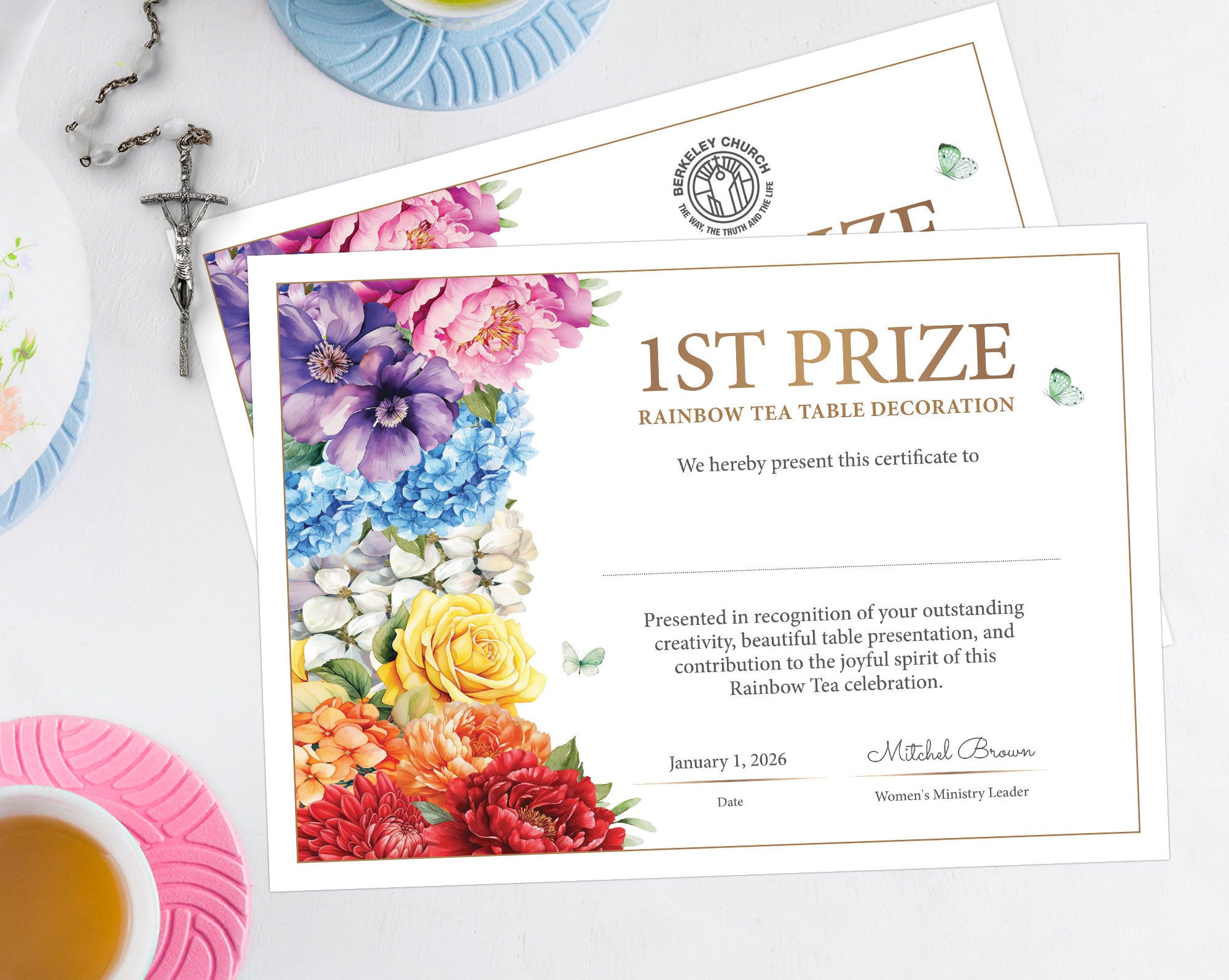 Two A4 printed certificates for 1st Prize Rainbow Tea Table Decoration, featuring a colorful floral border and decorative text, styled on a white tabletop with a teacup, saucer, cross, and plates in the background.