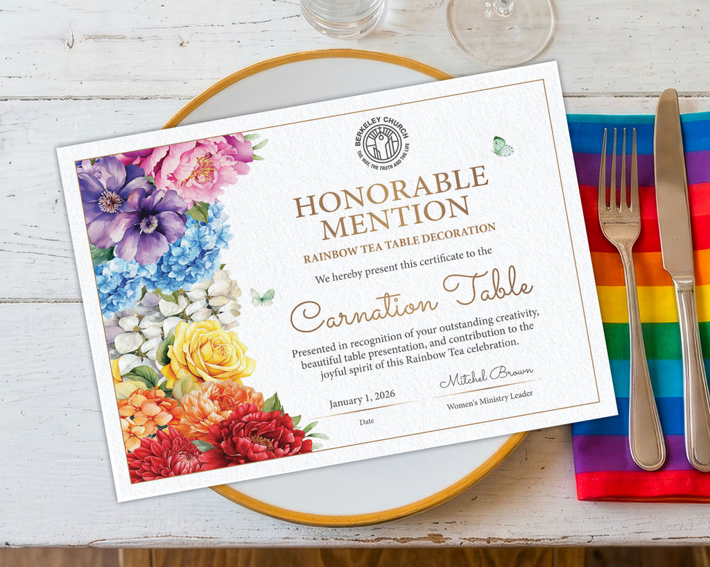 A printed A4 certificate card for a Rainbow Tea Table decoration sits on a gold-rimmed plate over a white wood surface, with a rainbow-striped napkin, silver fork, and knife beside it. The certificate features a floral border in purple, blue, yellow, orange, and red, with gold text reading 'Honorable Mention' and 'Rainbow Tea Table Decoration' and 'Carnation Table.