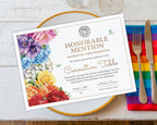 A printed A4 certificate card for a Rainbow Tea Table decoration sits on a gold-rimmed plate over a white wood surface, with a rainbow-striped napkin, silver fork, and knife beside it. The certificate features a floral border in purple, blue, yellow, orange, and red, with gold text reading 'Honorable Mention' and 'Rainbow Tea Table Decoration' and 'Carnation Table.