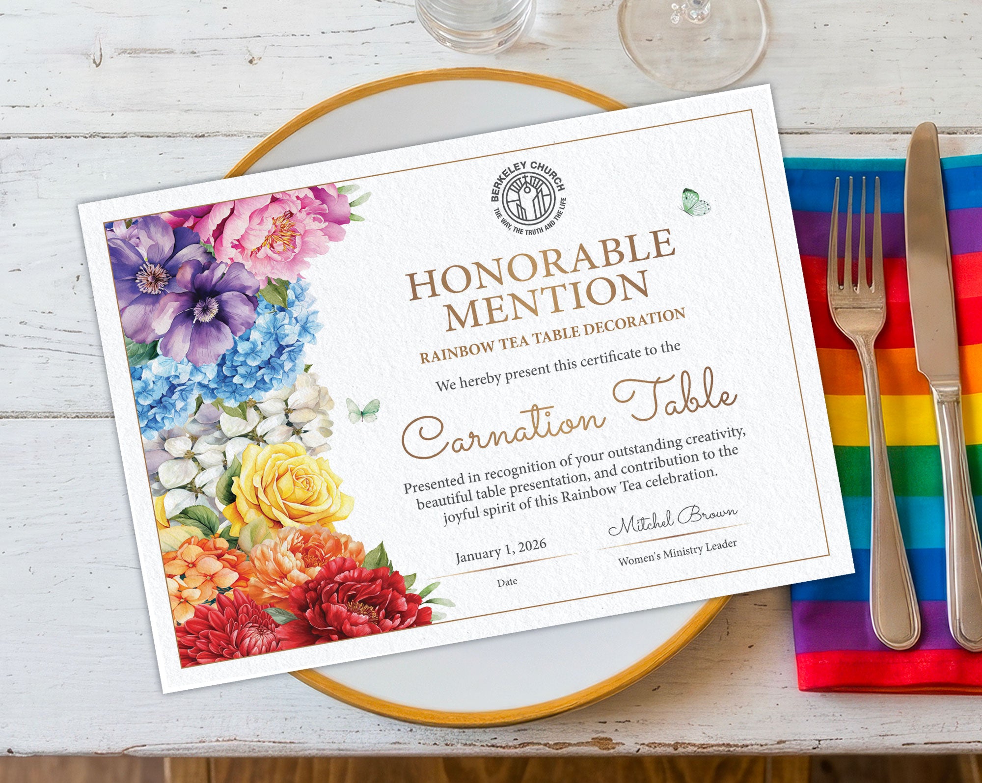 A printed A4 certificate card for a Rainbow Tea Table decoration sits on a gold-rimmed plate over a white wood surface, with a rainbow-striped napkin, silver fork, and knife beside it. The certificate features a floral border in purple, blue, yellow, orange, and red, with gold text reading 'Honorable Mention' and 'Rainbow Tea Table Decoration' and 'Carnation Table.