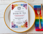 A printed fundraiser program card titled 'Rainbow Tea' sits centered on a white plate with a gold rim, alongside a rainbow-striped napkin, silver fork, and knife on a white wood tabletop.