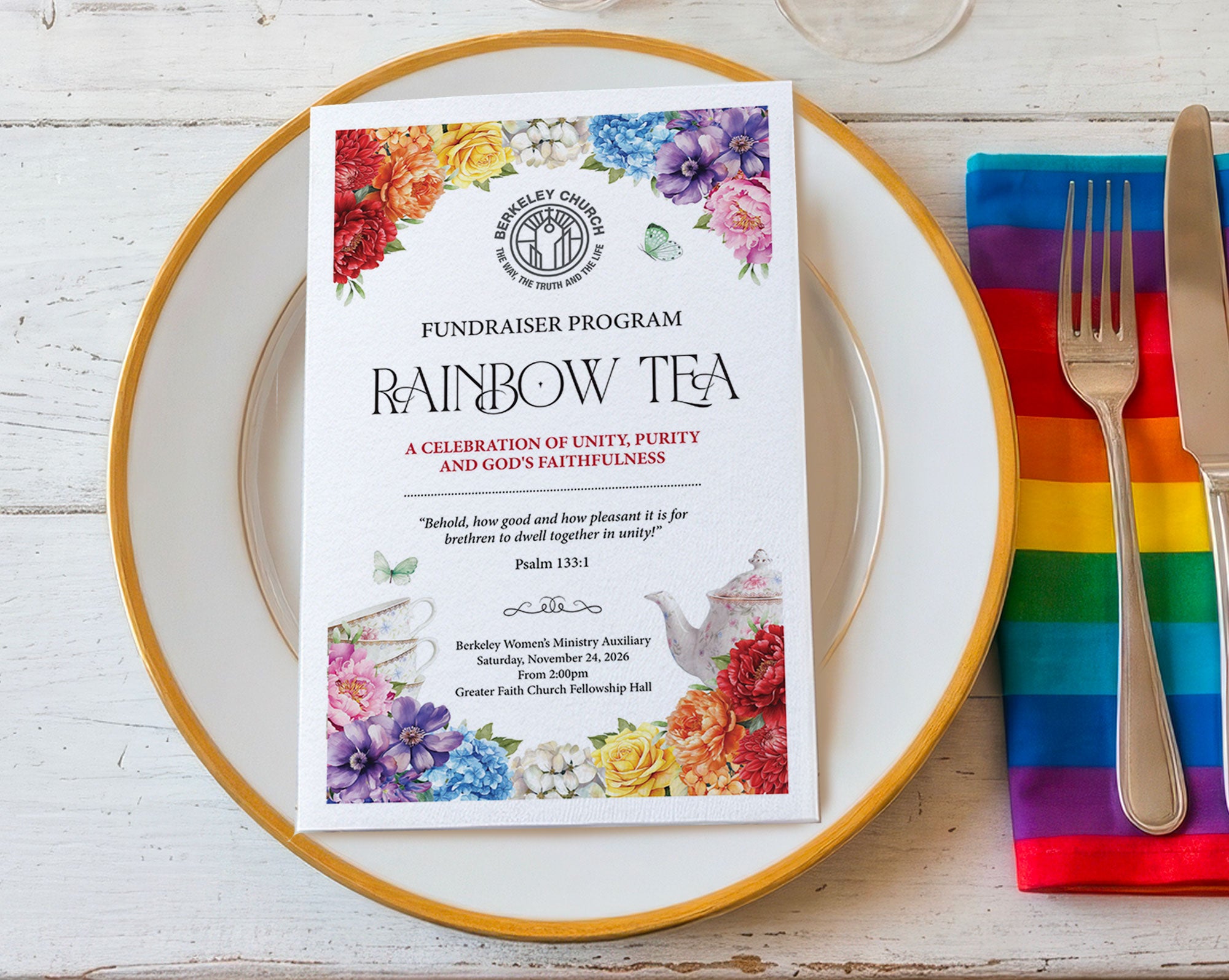 A printed fundraiser program card titled 'Rainbow Tea' sits centered on a white plate with a gold rim, alongside a rainbow-striped napkin, silver fork, and knife on a white wood tabletop.