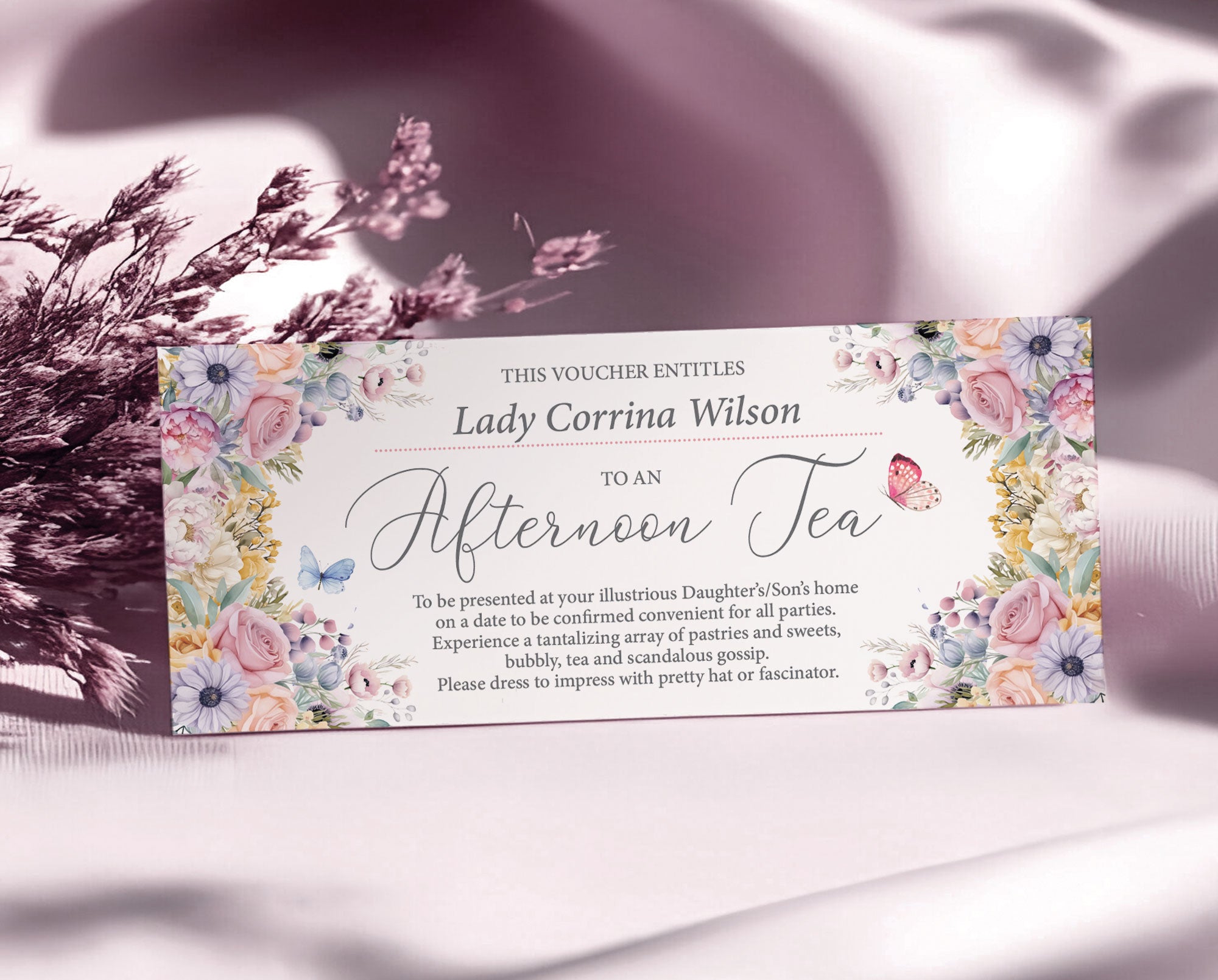 elegant pastel flower voucher for an afternoon tea with butterflies and text, leaning up against some dried flowers on a silk pink cloth