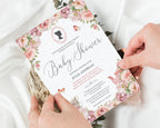 two hands holding a 5 x 7 blush pink regency style baby shower tea invitation over a white silk background