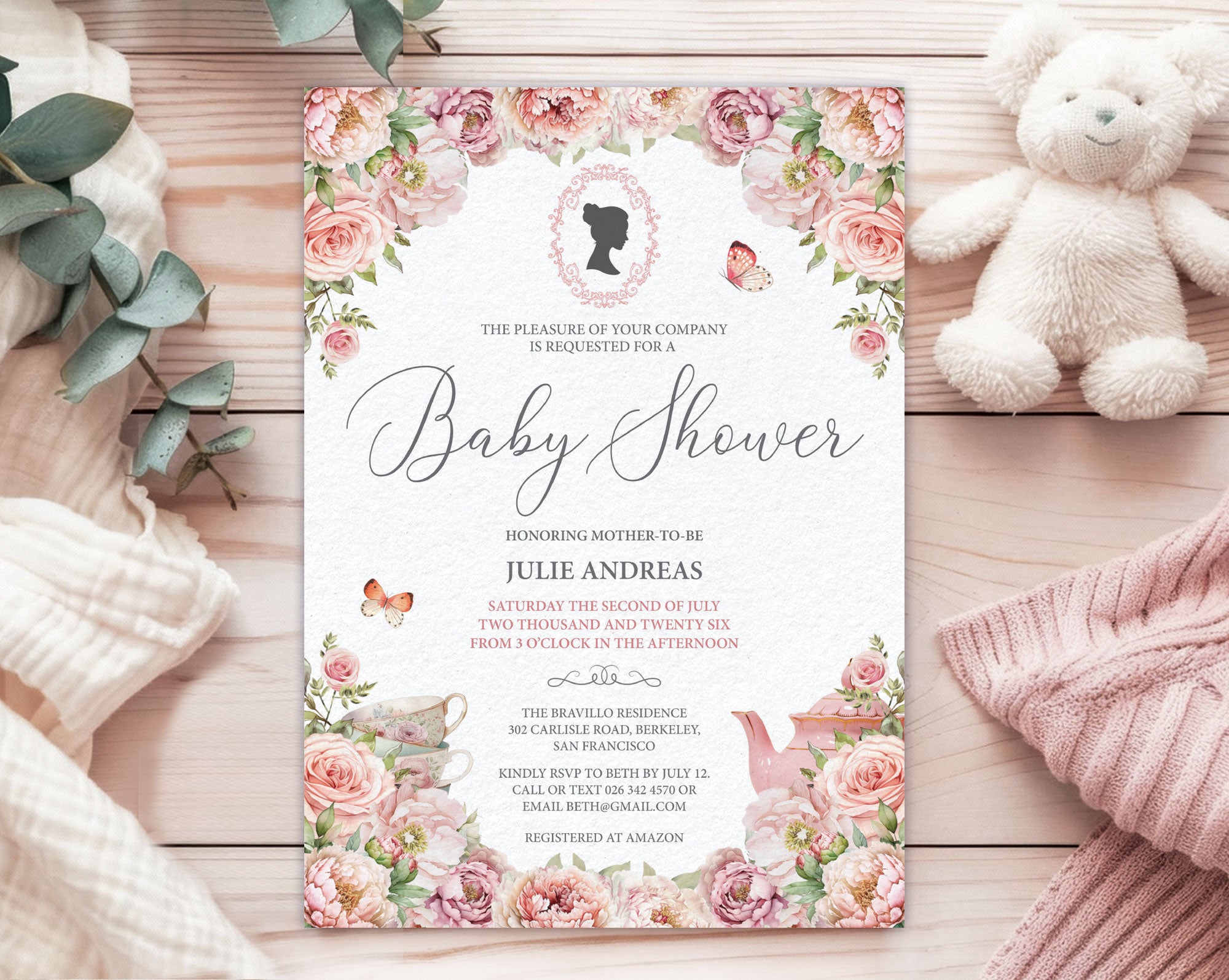 a blush pink flower baby shower tea invitation with cameo and teapot, laying on a wooden background next to a white teddy bear, some baby blankets and eucalyptus