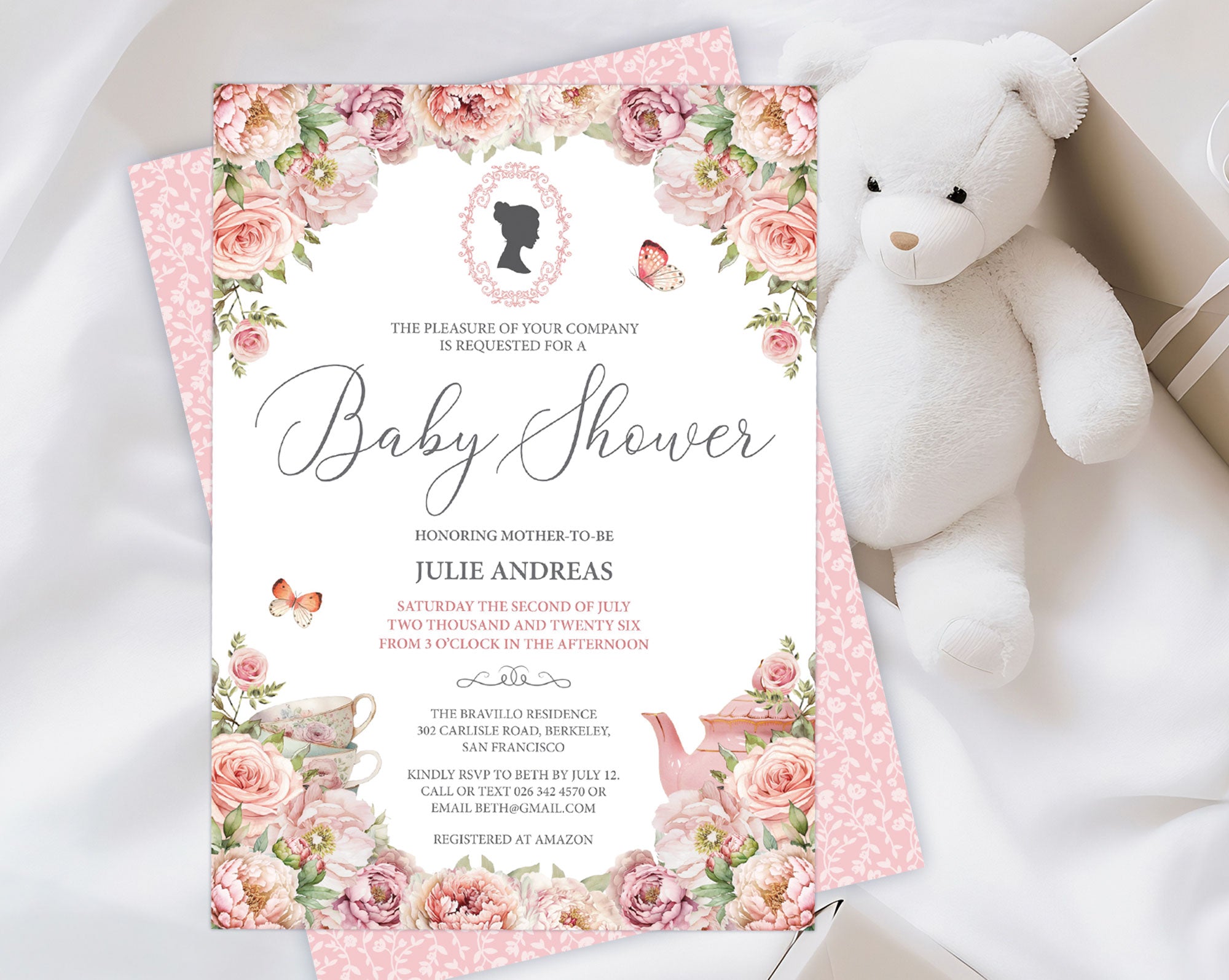 an elegant pastel baby shower tea regency style invitation laying on the pink pattern back of the invite, on a white cloth with a white teddy bear and a wrapped gift
