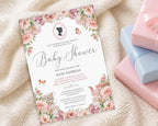 a blush pink baby shower invitation with cameo and flowers on a cream baby blanket next to some pink and blue wrapped gifts